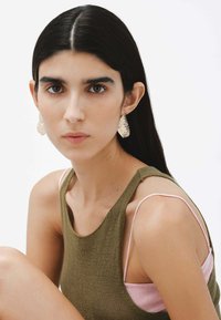 Pendientes texturizados en oro, top de punto verde oliva y tirantes rosa claro. Fondo neutro, centrado en los detalles de los accesorios y la prenda.