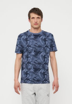 Férfi kék mintás rövid ujjú pólóban és világosszürke melegítőnadrágban, sima fehér háttér előtt áll.