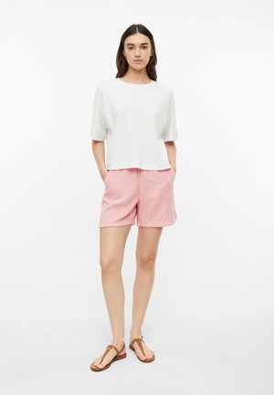 Woman standing with hands in pockets wearing white short-sleeve top, pink and white striped shorts, and brown sandals on white background.