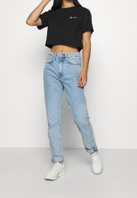 Camiseta negra corta con un logo colorido, jeans de talle alto en azul claro y zapatillas blancas. Conjunto exhibido en un modelo de pie.