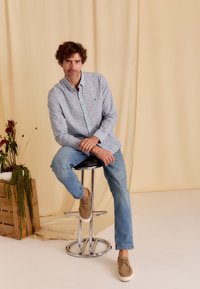 Lichtblauw patroon shirt met een knoopsluiting, gecombineerd met lichte spijkerbroek en beige schoenen. Achtergrond is voorzien van zachte gele draperieën.