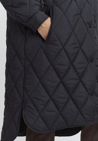 Manteau long matelassé bleu marine, avec des motifs en losange, des boutons-pression et des poches latérales, un ourlet courbé et une texture douce.
