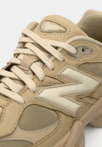 Baskets beige New Balance 9060 avec des lacets épais texturés, des panneaux en daim et en mesh, et un grand logo blanc "N" sur le côté.