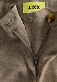 Bruine broek met gedeeltelijk open ritssluiting, knoop dichtgemaakt, en een geel label met de tekst "JJXX" aan de binnenkant van de tailleband.