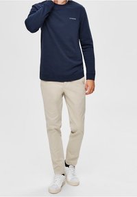 Námořnická mikina s dlouhým rukávem, kulatý výstřih, malý bílý text na hrudi, spárovaná se světle béžovými kalhotami a bílými teniskami. Hladká textura látky.