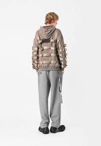 Sudadera marrón con tiras beiges en capas en la parte posterior, combinada con pantalones de chándal grises y calzado negro texturizado. Diseño minimalista.