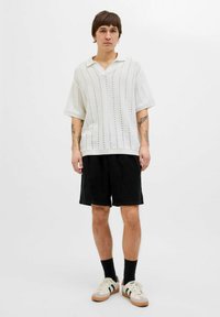 Hvid strikket polo med korte ærmer og v-hals, parret med sorte knælange shorts og beige sneakers, der har grønne striber.