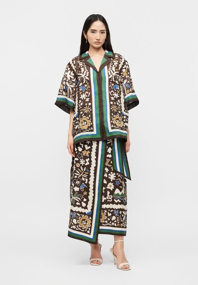 Γυναίκα που φοράει χαλαρή, καφέ σετ δύο τεμαχίων με μοτίβα και μπλε, πράσινες και λευκές πινελιές, στέκεται με ουδέτερη έκφραση και λευκά τακούνια.