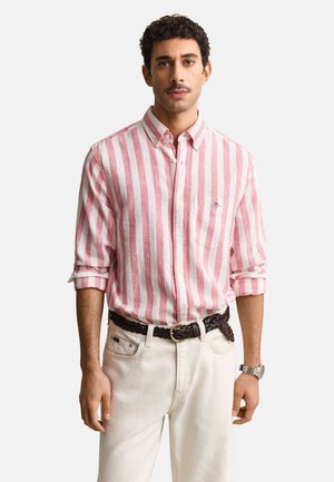 Mann trägt ein rosa-weiß vertikal gestreiftes Hemd mit Knöpfen, weiße Hose mit geflochtenem braunem Gürtel und eine silberne Armbanduhr, steht frontal.