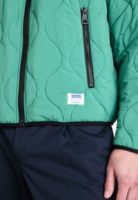 Quilted grøn jakke med en sort lynlås, strukturerede bølge-mønstre og et blåt mærke med "HUGO." Kantet i sort. Mørkeblå bukser synlige.