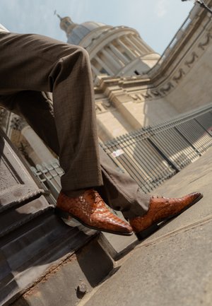 Person iført brune flettede lædersko og brune bukser sidder på en stenskammel nær en historisk bygning med søjler og kuppel.