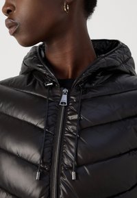 Veste doudoune noire avec capuche, présentant une texture matelassée, une fermeture éclair et des cordons de serrage réglables pour un ajustement parfait.