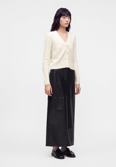 Femme portant un cardigan crème à col en V avec des nœuds, une longue jupe en cuir noire et des mocassins noirs, debout devant un fond clair uni.