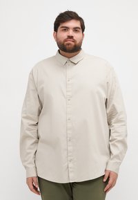 Camisa beige de manga larga con cuello abotonado, que presenta un corte relajado y una textura suave, combinada con pantalones verde oliva.