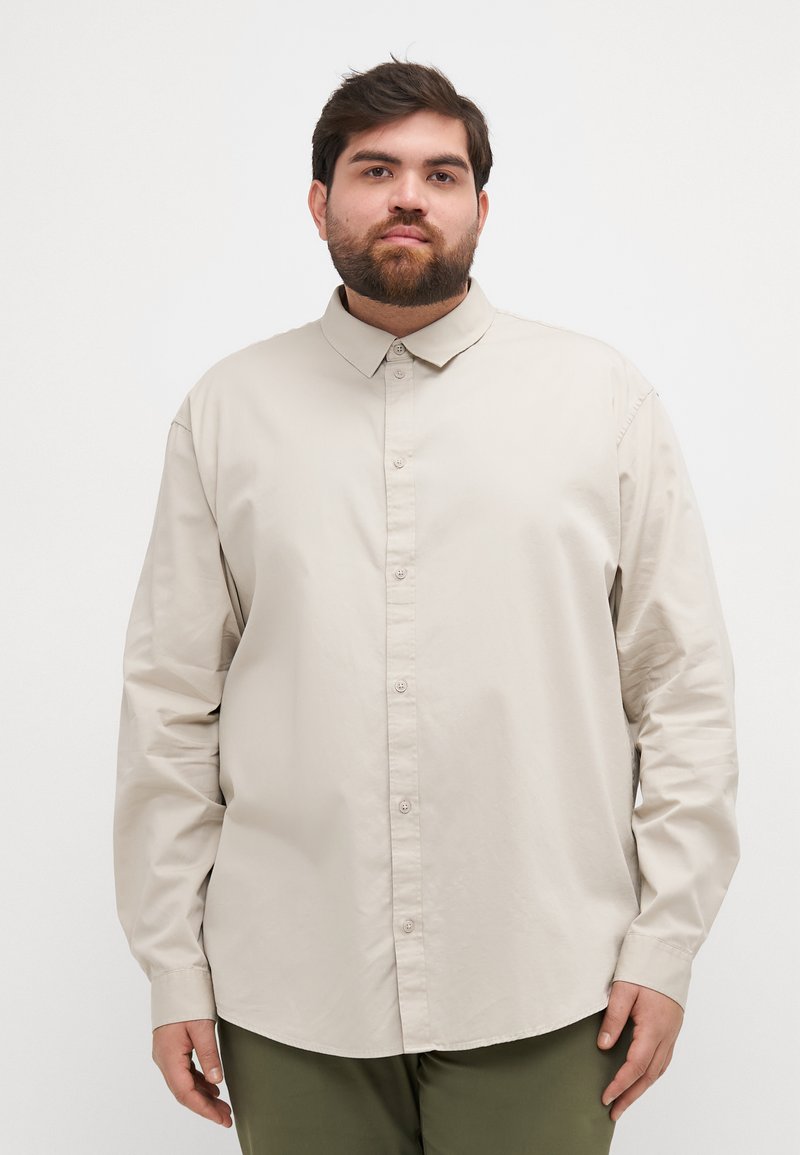 Camisa beige de manga larga con cuello abotonado, que presenta un corte relajado y una textura suave, combinada con pantalones verde oliva.