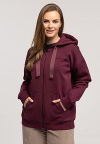 Burgundowa bluza z kapturem na zamek, wykonana z miękkiej bawełny z dodatkiem syntetyków, z przestronnym kapturem, przednimi kieszeniami oraz markowymi sznurkami. Gładka textura.