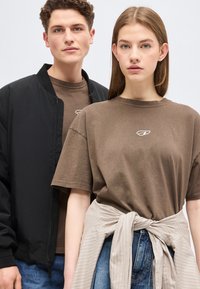 Νεαρός άνδρας και γυναίκα που φορούν καφέ μπλουζάκια με μικρό λευκό λογότυπο, η γυναίκα έχει ριγέ πουκάμισο δεμένο γύρω από τη μέση, στέκονται μπροστά σε απλό φόντο.