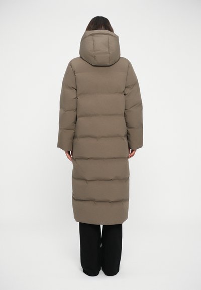 Manteau long rembourré en vert olive avec une capuche, doté d'un design matelassé et d'une silhouette droite. Porté sur un pantalon noir.
