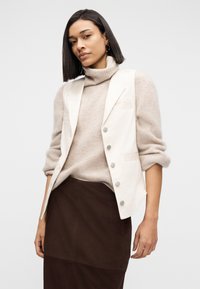 Femme aux cheveux longs et lisses, portant un pull à col roulé beige, un gilet blanc boutonné, des boucles d'oreilles dorées en créole et une jupe marron foncé, sur fond blanc.