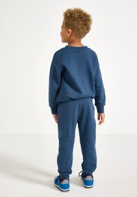 Lapsi, jolla on kiharaiset hiukset, seisoo selin sinisessä hupparissa, vastaavissa housuissa ja sinisissä lenkkareissa, joissa on violetteja ja harmaita yksityiskohtia.