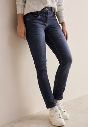 Dark blue jeans with a slim fit, featuring contrast stitching, front pockets, and a button closure, paired with white sneakers.