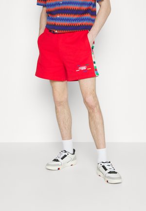 Κόκκινες βαμβακερές βερμούδες με σχέδιο με λογότυπο, πλαϊνές ρίγες σε διάφορα χρώματα και τυπική εφαρμογή. Συνδυάζονται με λευκές κάλτσες και αθλητικά παπούτσια.