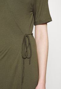 Gros plan sur une robe vert olive à manches courtes avec un cordon de serrage noué sur le côté, porté sur un bras à peau claire, sur un fond neutre.
