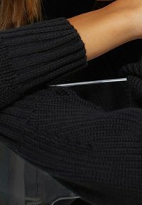 Czarny sweter dziany o prążkowanej fakturze, prezentujący zbliżenie na rękaw i ramię, z eleganckim, przezroczystym detalem akcesorium.