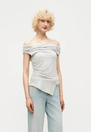 Γυναίκα με κοντά σγουρά ξανθά μαλλιά που φορούσε ανοιχτό γκρι τοπ με ρυτιδωτό σχέδιο και ανοιχτό μπλε τζιν, στέκεται μπροστά σε απλό φόντο.