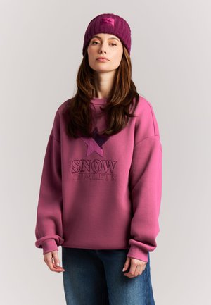 Woman wearing a pink beanie and pink sweatshirt with "SNOW MUCH FUN" text and star graphic, paired with blue jeans, standing against plain background.