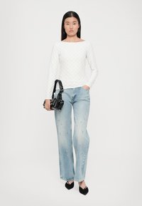 Hvit langermet topp med et teksturert diamantmønster, kombinert med lyseblå vide jeans med dekorasjoner, og svarte spisse sko.