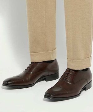 Brown leather dress shoes with laces worn with beige trousers, shown from the lower leg to foot against a plain background.
