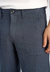 Mörkblå herringbone-mönstrade byxor med knappstängning, femfickorsdesign och textilstruktur, som visar midjebandet och sidofickan.