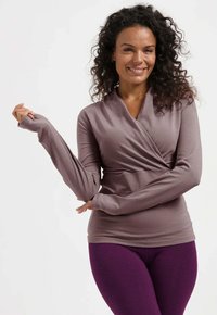 Langärmliges, wickelförmiges Oberteil aus leicht mauvefarbener Stoff, mit V-Ausschnitt und taillierter Passform, kombiniert mit dunkelvioletten Leggings.