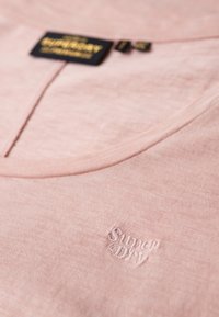 Różowa bawełniana koszulka z dekoltem w serek, z subtelnym, tonowanym haftowanym logo "Superdry" z przodu. Gładka faktura.