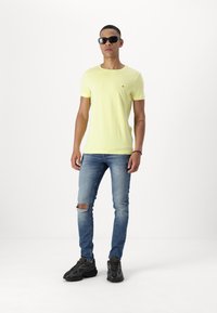 Tricou galben cu mânecă scurtă, având un mic logo, asortat cu blugi deschiși, rupti, și adidași negri. Ochelarii de soare completează look-ul.
