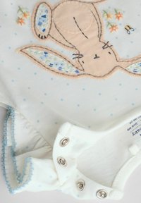 Bomulds baby bodystocking med en kaninbrodering, lyse pastelfarver, små blå prikker og trykknaplukninger med en flæsekant.