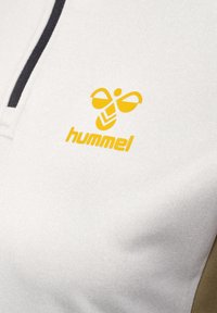 Biała sportowa bluzka z zapięciem pod szyję, czarnymi akcentami i żółtym logo "hummel" z motywem pszczoły na piersi. Gładka faktura materiału.