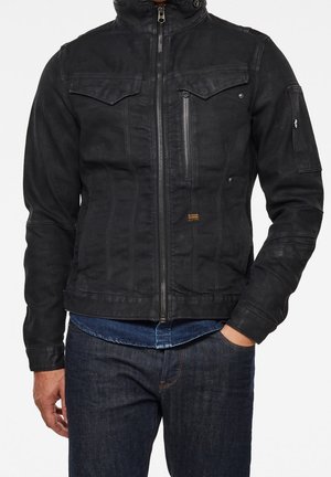 Homme portant une veste noire zippée avec des poches poitrine et une poche zippée, associée à un jean bleu foncé et une chemise bleue en dessous.