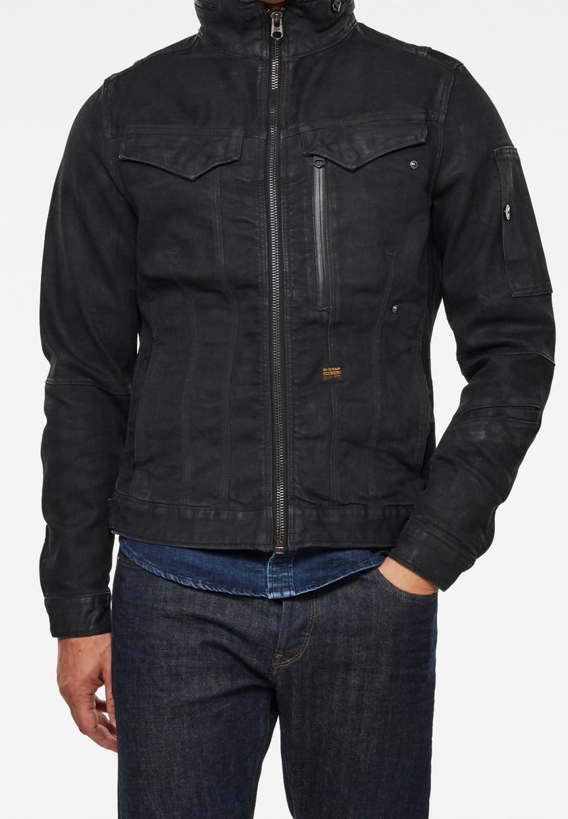 Homme portant une veste noire zippée avec des poches poitrine et une poche zippée, associée à un jean bleu foncé et une chemise bleue en dessous.