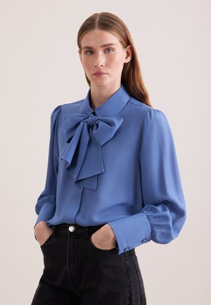 Donna con capelli lunghi castani, che indossa una camicetta blu con un grande fiocco e jeans neri, mani nelle tasche, sfondo neutro.