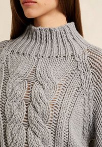 Femme portant un pull en maille épaisse gris clair avec des motifs torsadés et un col montant côtelé, montrant le cou et le bas du visage.