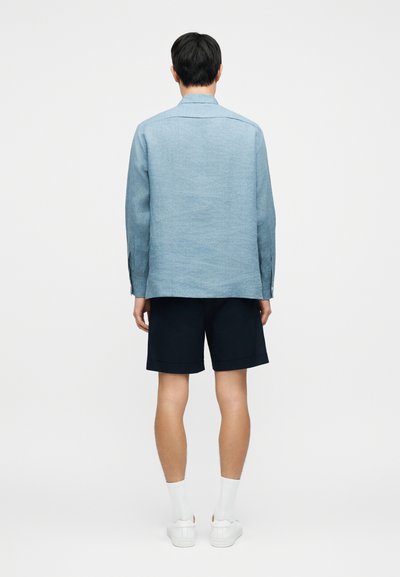 Ανοιχτό μπλε πουκάμισο με κουμπιά, μακριά μανίκια και ίσια γραμμή, συνδυασμένο με σκούρα μπλε σορτς. Λευκές κάλτσες και αθλητικά παπούτσια ολοκληρώνουν το σύνολο.