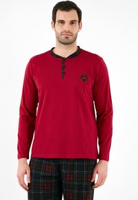 Rød langærmet skjorte med en sort og ternet krave, der har tre knapper og et rundt logo. Båret sammen med sorte ternede pyjamasbukser.