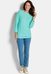 Türkisfarbenem Strickpullover mit weitem Rundhalsausschnitt, langen Ärmeln, kombiniert mit hellblauen Jeans und beigen Sneakers. Weiche Textur, lockere Passform.