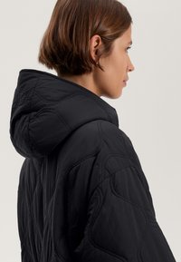 Veste matelassée noire avec capuche, présentant une texture douce et un motif de coutures géométriques. Comprend une coupe décontractée et un tissu lisse.