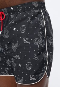 Μαύρα μαγιό σορτς με λευκά σχέδια εμπνευσμένα από τατουάζ, που περιλαμβάνουν φίδια, λουλούδια, πουλιά και τράπουλες με την επιγραφή «Καλή Τύχη», με κόκκινο κορδόνι στο πλάι.