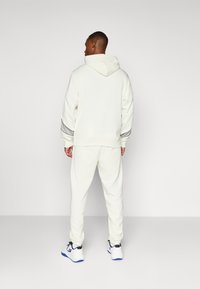 Sudadera blanca con capucha y pantalones a juego, fabricados con tejido suave. Presenta un diseño sencillo con mangas y tobillos en puños, combinados con zapatillas blancas.
