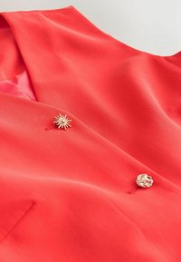 Punane bleiser kahe dekoratiivse kuldse nupuga, üks päikese kujuline ja teine abstraktse tekstuuriga.