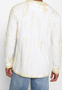 White long-sleeve shirt with yellow tie-dye pattern, ribbed cuffs, and a relaxed fit, paired with light blue denim jeans.